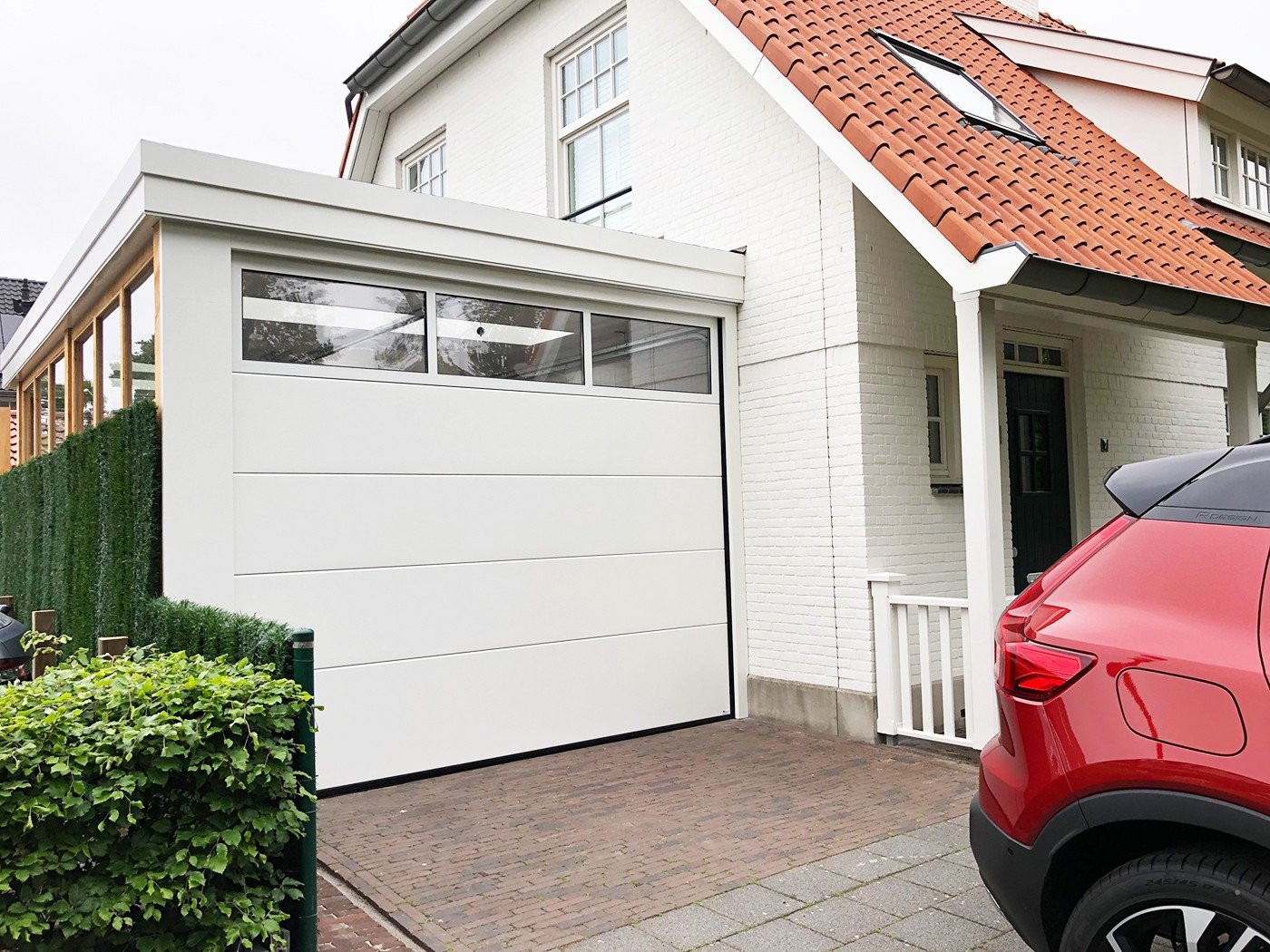 Moderne sectionaalpoort in een stijlvolle, witte afwerking bij een woning in Sittard, met een groenhekje en rode auto vooraan.