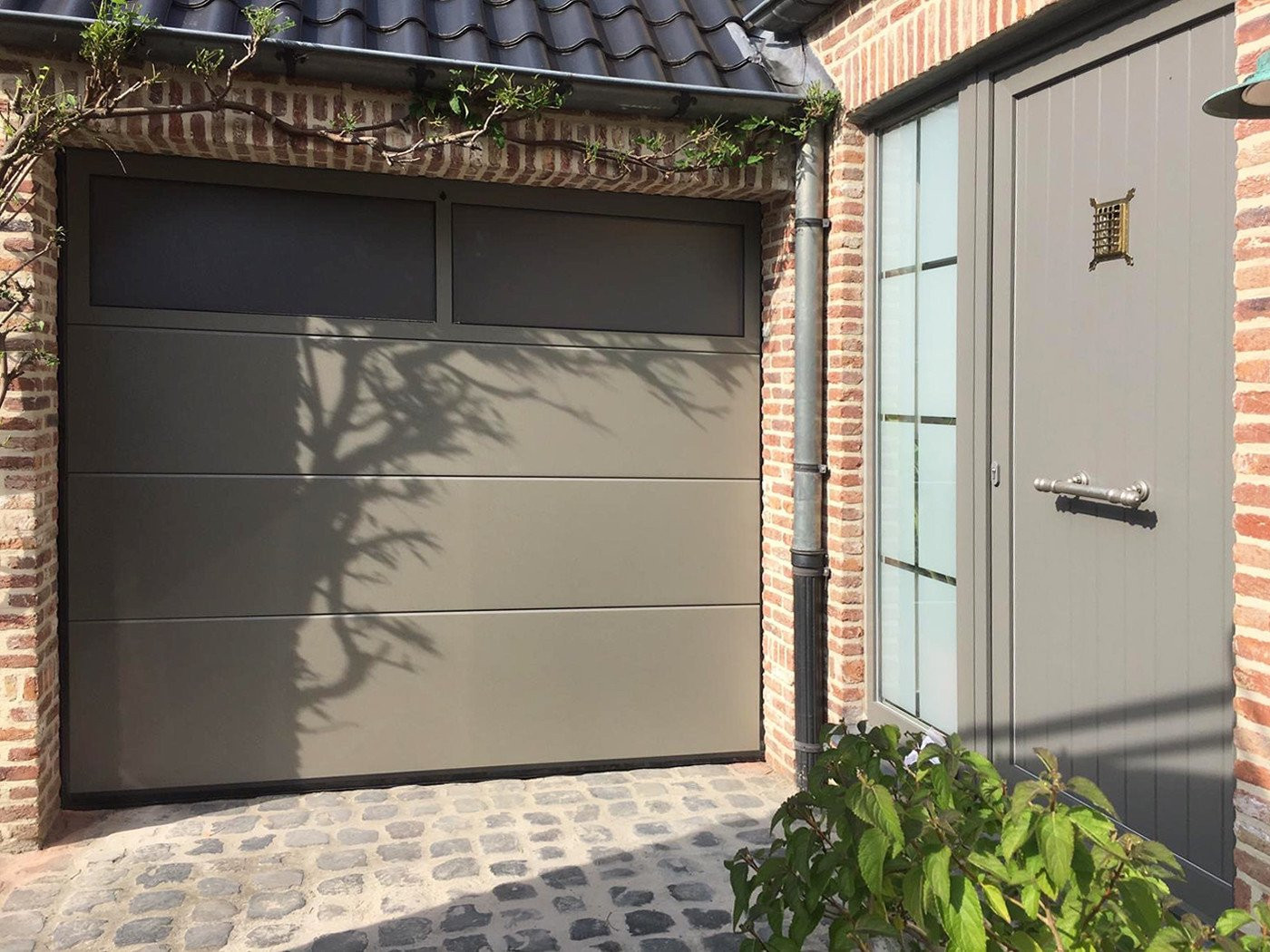 Moderne sectionaalpoort in neutrale tint, passend bij een bakstenen muur en entree, met subtiele schaduw van planten.