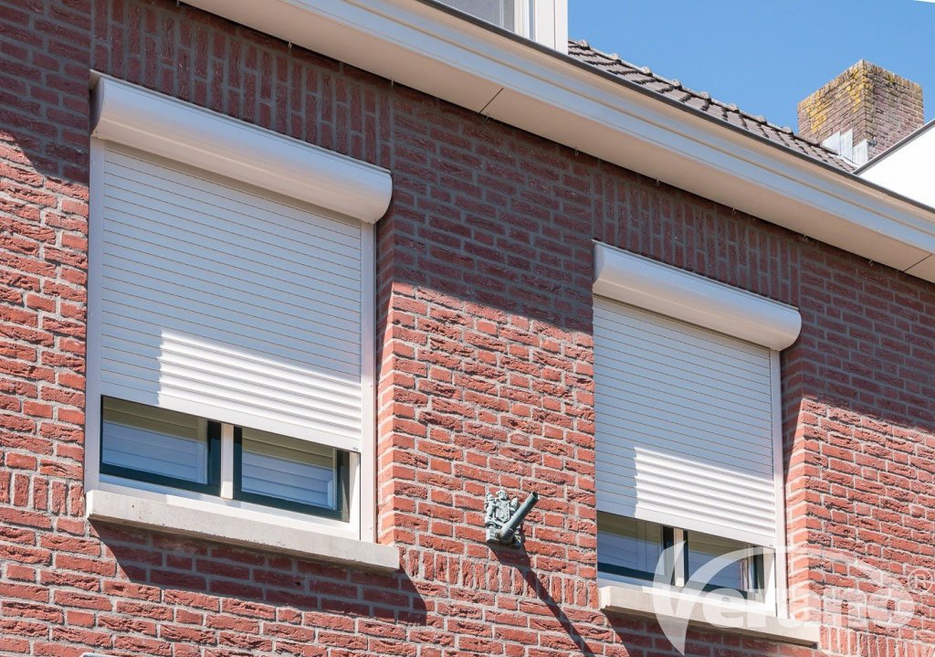 Hoogwaardige handbediende rolluiken voor uw woning. Handbediende rolluiken in een lichte kleur, geplaatst op een bakstenen gevel, ideaal voor privacy en zonwering.