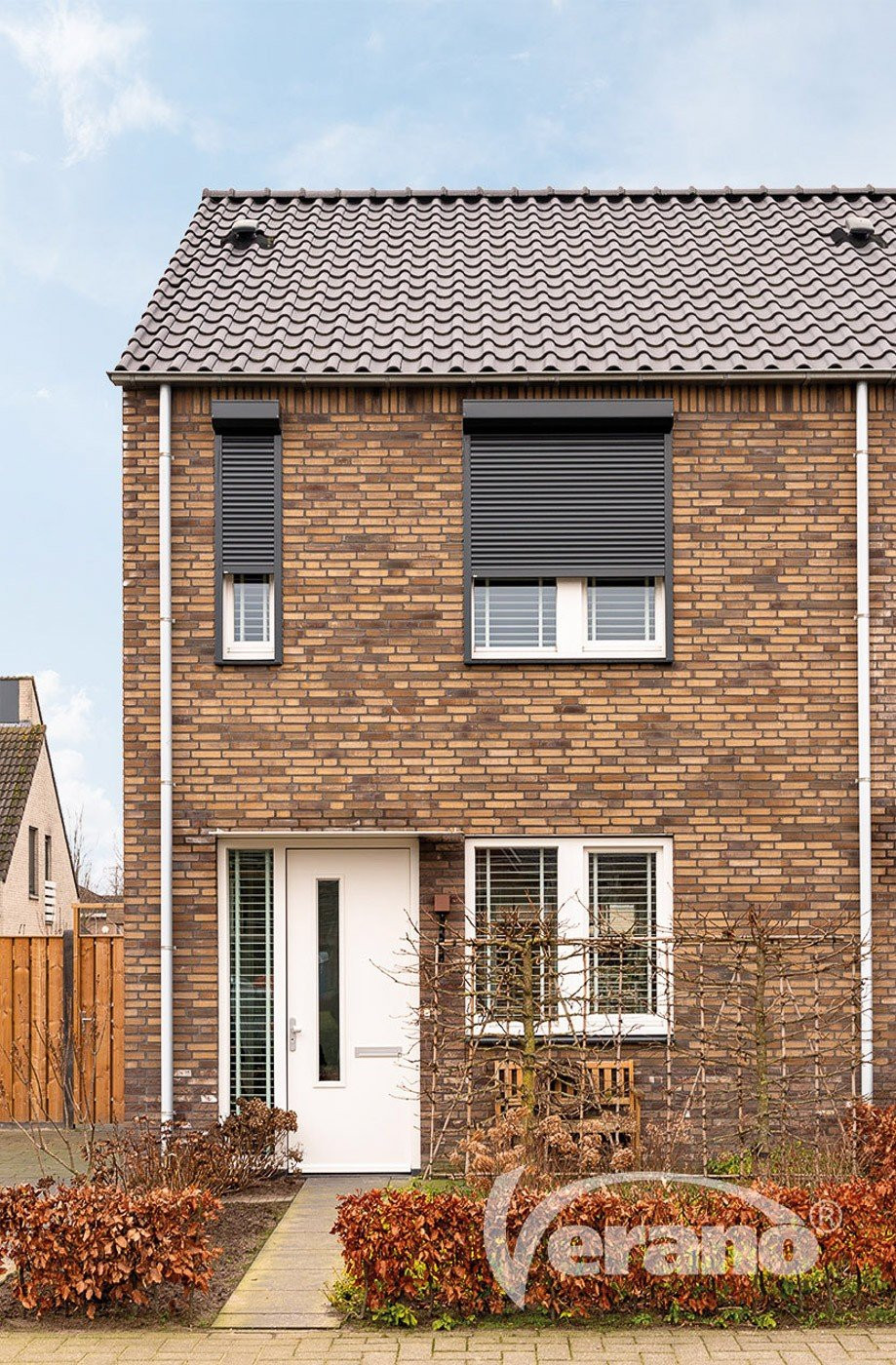 Stijlvolle handbediende rolluiken voor uw thuis! Handbediende rolluiken in zwart boven een woning met bakstenen gevel.