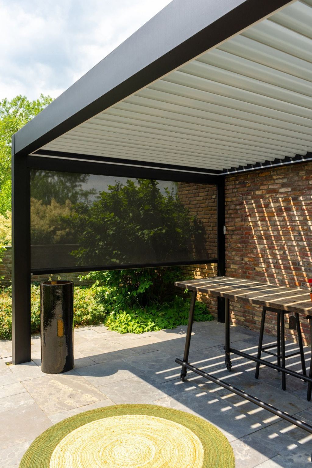 Black patio cover with a louvred roof with white louvres, finished with screens, an attractive bar table and a green rug.