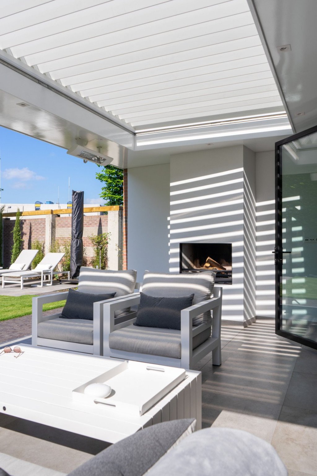 Ook genieten van een overkapping met lamellendak? Renoco monteert in Sittard - Geleen en Limburg!. Moderne veranda met lamellendak met witte lamellen over een elegante zithoek en strak terras, met comfortabele tuinmeubels.