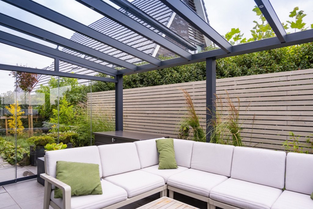 Prachtige terrasoverkapping voor uw huis in Sittard - Geleen of elders in Limburg. Moderne terrasoverkapping aan huis met glazen schuifwanden, omringd door groen, en strakke tuinmeubelen.