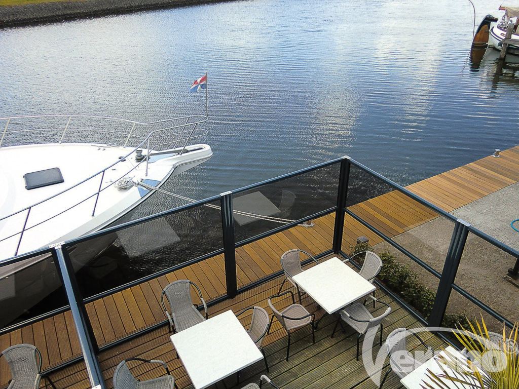 Glazen windscherm scheidt terras van water, met gezellige tuinmeubels en een jacht.