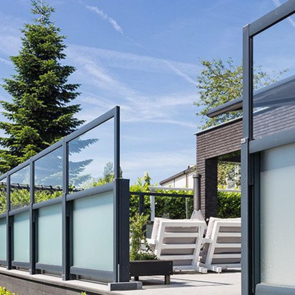 Eleganter Windschutz aus Glas trennt die Terrasse vom Garten, mit modernen Gartenm&ouml;beln auf der Terrasse.