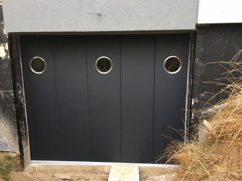 Modern side-sliding sectional door with round windows in a sleek black finish. Suitable for any type of home.