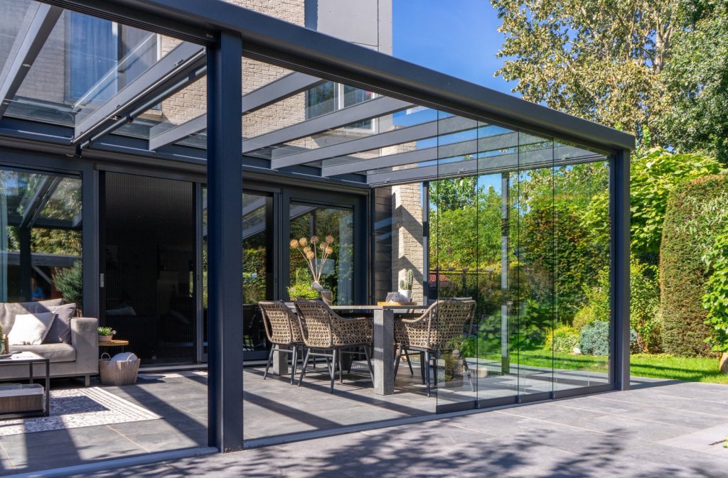 Glazen schuifwanden kopen? Renoco monteert in Sittard - Geleen Limburg. Strakke terrasoverkapping met glazen schuifwanden, moderne tuinmeubelen in een mooie tuin.