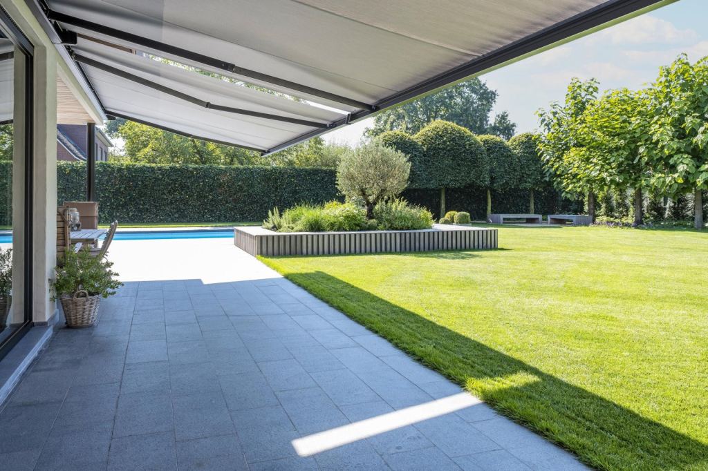 Folding-arm awning over a patio with a green garden and swimming pool in the background.