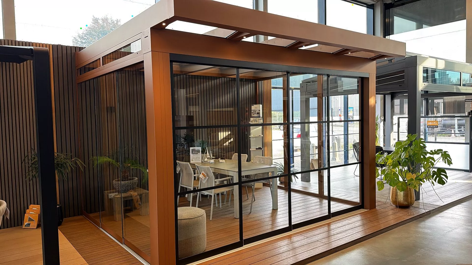 Wood-coloured patio cover with sliding glass walls and a dining table in the Renoco showroom