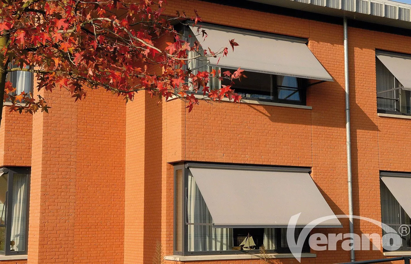 Graue Fallarmmarkisen an den Fenstern eines orangefarbenen Gebäudes mit blaugrauen Fensterrahmen.