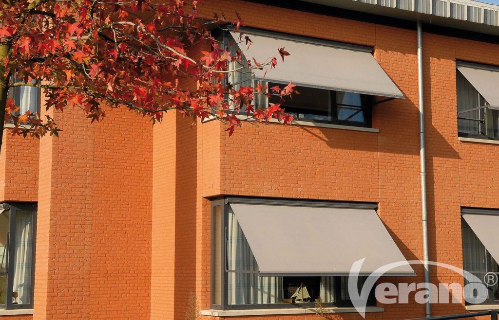 Graue Fallarmmarkisen an den Fenstern eines orangefarbenen Geb&auml;udes mit blaugrauen Fensterrahmen.
